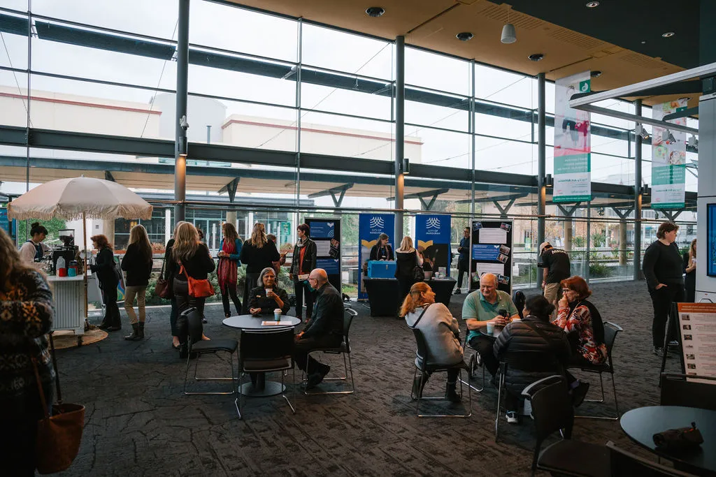 People gathered in a large, bright lobby space, talking around tables and visiting display stands.